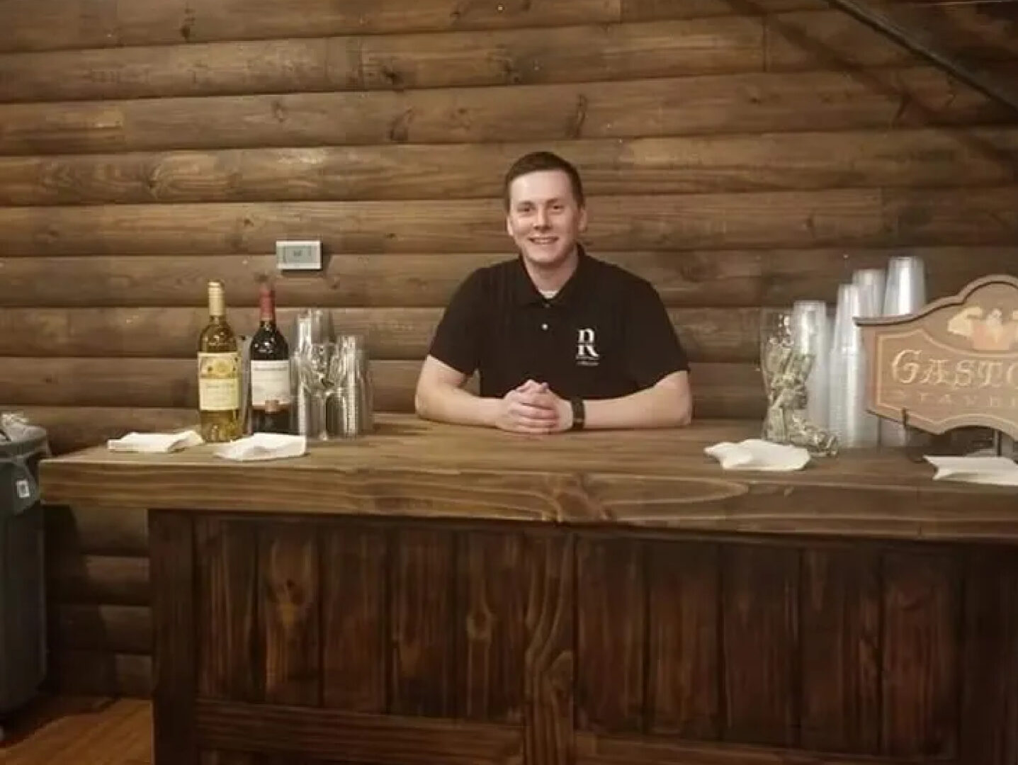 Man wearing a black polo shirt standing behind a wooden bar table in a cabin-like room