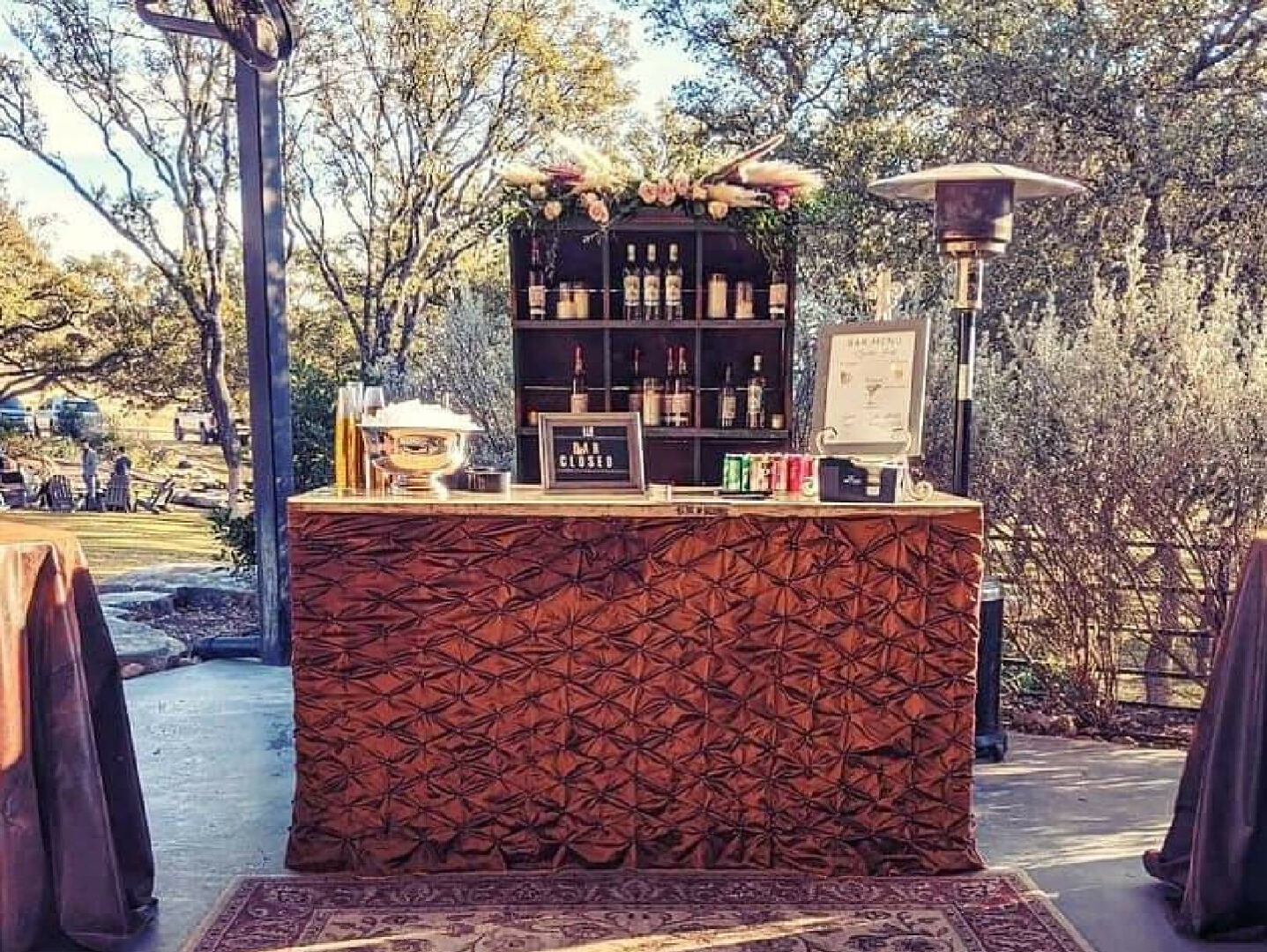 Decked-out bar table fitted with a ribbed table cloth in front of an alcohol cabinet placed in an open environment